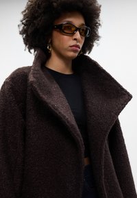 Abrigo marrón texturizado con un amplio cuello y un corte oversize, superpuesto a una blusa negra ajustada. Presenta un material suave y peludo con detalles sutiles.