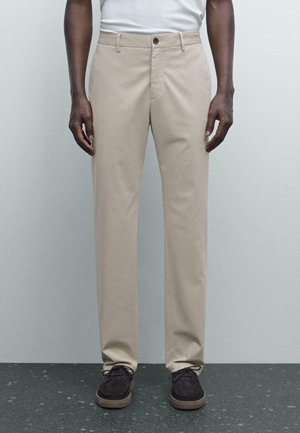 Uomo con pantaloni beige a gamba dritta e scarpe marroni scure con lacci, in piedi contro un muro grigio chiaro e un pavimento scuro macchiato.