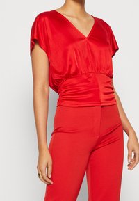 Rød satin top med V-udskæring og korte ærmer, med rynkede detaljer ved taljen, kombineret med matchende røde, tætsiddende bukser.