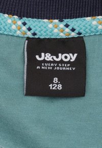 Label bevat een zwarte tag met witte tekst, waarop staat "J&JOY EVERY STEP A NEW JOURNEY," maat "8," en "128." De elastische tailleband is turkoois met geweven accenten.