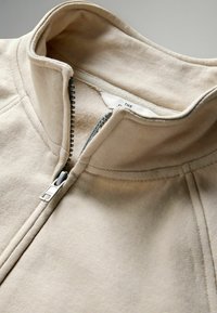 Chaqueta beige con cremallera y cuello alto, fabricada en una suave mezcla de algodón, que presenta una cremallera metálica resistente y detalles de costura visibles.