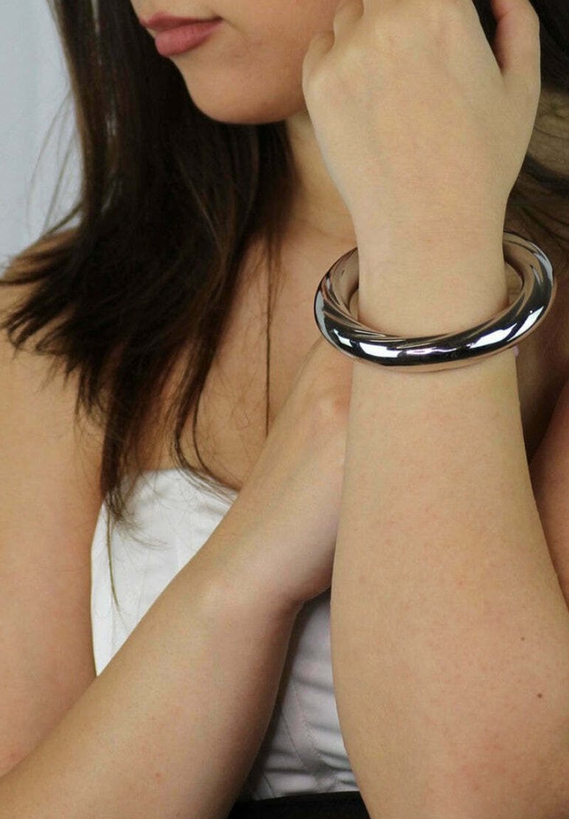 Srebrna metalowa bransoleta w kształcie bangle z gładką, błyszczącą powierzchnią. O okrągłym kształcie, bezszwowa konstrukcja, noszona na nadgarstku osoby.