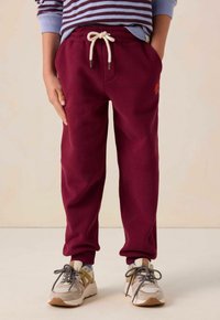 Bordeauxrote Sweatpants mit einem Kordelzugbund, elastischen Bündchen und seitlichen Taschen. Kombiniert mit grauen und beigen Sneakers.