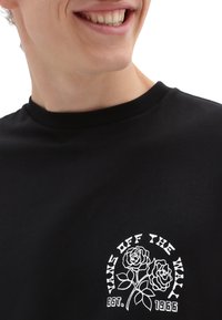 Musta puuvillapaita, jossa on valkoinen grafiikka kahdesta ruususta ja teksti "VANS OFF THE WALL EST. 1966" ympyrämuotoisessa suunnittelussa.