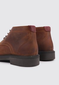 Botines de cuero marrón con punta redondeada, suela de goma negra y detalle en el tacón color burdeos. Textura lisa con detalles de costura.