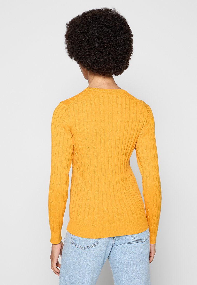 Żółty sweter w splot kablowy z długimi rękawami, ściągaczem u dołu i dopasowanym krojem, zestawiony z jasnoniebieskimi dżinsami.