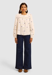 Camicia bianca con ricami floreali blu, abbinata a jeans blu a gamba larga e scarpe in suede marroni. Maniche lunghe raccolte ai polsi.