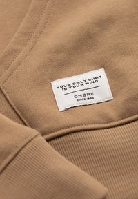 Hnědá mikina z jemného materiálu s měkkou strukturou, na které je bílá nálepka s textem "TVOJÍ JEDINOU MEZÍ JE TVÁ MYSL" a "OMBRA od 1985."