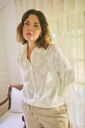Femme aux cheveux bruns mi-longs portant un chemisier blanc à motifs et un pantalon beige, debout à l'intérieur près d'une fenêtre avec des voilages.