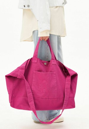 Persoon draagt lichte spijkerbroek en witte jas en houdt een grote felroze draagtas met voorvak en knoopsluiting vast.