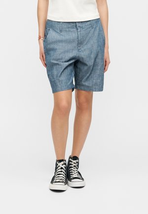 Persona vistiendo pantalones cortos azules hasta la rodilla, camiseta blanca, zapatillas negras altas, de pie con las manos en los bolsillos contra un fondo blanco.