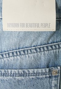 Vaaleansininen denim-kangas, jossa on teksturoitu pinta. Ominaisuuksiin kuuluu tikattu tasku ja valkoinen nahkalappu, jossa on kohokuvioituna "DRYKORN FOR BEAUTIFUL PEOPLE".