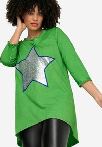 Tunique en coton verte avec un design de star argenté brillant entouré de bleu. Dotée de manches trois-quarts et d'un ourlet arrière plus long.