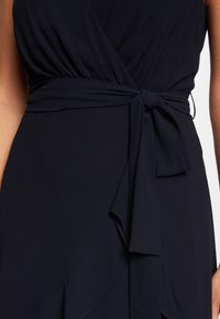 Navyblaues Kleid mit Wickeldesign, ausgestattet mit einem gebundenen Taillenbund und fließendem Stoff. Glatte Textur mit sanften Drapierungen und leichtem Rüscheneffekt.