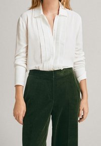 White blouse with textured vertical pleats, collared neckline, and long sleeves, paired with dark green corduroy pants.