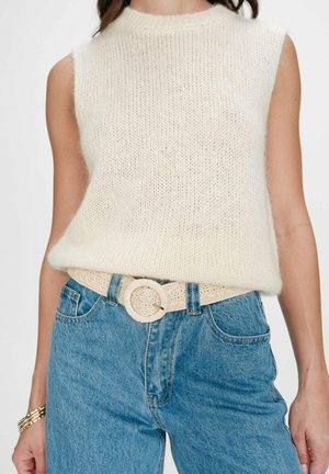 Femme portant un haut sans manches en maille blanche rentré dans un jean bleu taille haute avec une ceinture beige tissée et des bracelets en or au poignet gauche.