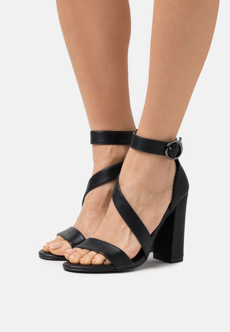 Pieds portant des sandales à talons hauts noires avec des brides de cheville et des fermetures à boucle ronde sur un fond clair.
