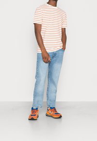 Hombre de pie con las manos en los bolsillos, vestido con una camiseta de rayas blancas y naranjas, jeans de mezclilla azul claro y zapatillas naranjas y azules sobre un suelo gris.