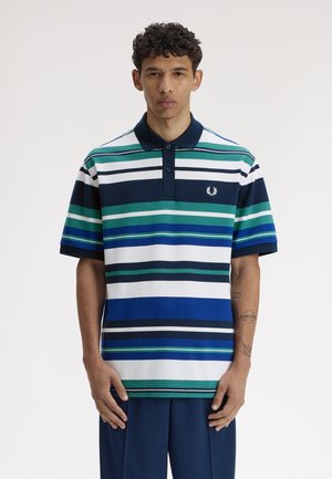 Homme portant un polo à manches courtes rayé bleu marine, vert, blanc et bleu avec col, debout devant un fond clair uni.