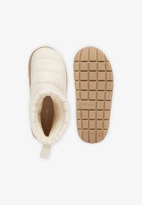 Botín sin cordones en color crema de Lacoste con parte superior acolchada, cuello acolchado y suela de goma texturizada marrón con patrón de rejilla para un mejor agarre.