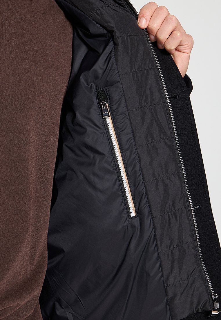 Intérieur d'une veste noire montrant une doublure matelassée, avec une poche zippée et une quincaillerie de fermeture éclair en argent, le tout avec une texture lisse et légèrement brillante.