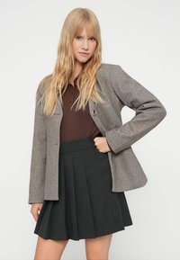 Blazer marrón de espiguilla con dos botones, llevado sobre una parte superior de canalé oscuro y una falda negra con pliegues, que muestra un diseño y textura estructurados.