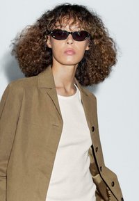 Giacca esterna marrone, top bianco e occhiali da sole scuri. La giacca presenta una texture liscia e grandi bottoni, mentre gli occhiali da sole hanno un design elegante e stretto.