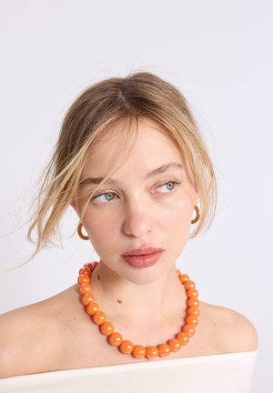 Mladá žena s blond vlasmi nosí veľký oranžový korálkový náhrdelník a zlaté kruhové náušnice, pozerá sa mierne do strany na bielom pozadí.