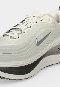 Λευκό και γκρι αθλητικό παπούτσι Nike με κυματιστό υφασμάτινο επάνω μέρος, ασημένιο λογότυπο Swoosh, λευκά κορδόνια και παχιά μαξιλαρωτή σόλα με μαύρη λεπτομέρεια.