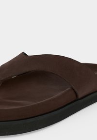 Καφέ δερμάτινο σανδάλι τύπου φλιπάρο με φαρδιά λουριά και μαύρη λαστιχένια σόλα, εμφανίζεται σε κοντινό πλάνο με λευκό φόντο.