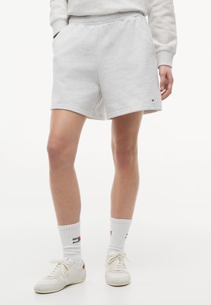 Osoba ubrana w jasnoszare szorty ze ściągaczem, białe skarpetki za kostkę z logo oraz białe sneakersy na sznurowadła, stojąca na białym tle.