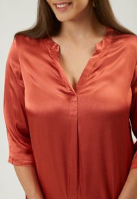 Seidenweiches, rostfarbendes Blusenshirt mit V-Ausschnitt, 3/4-Ärmeln und dezenter Ausstrahlung. Verfügt über eine Knopfleiste und eine glatte Textur.