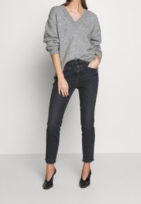 Suéter de punto gris con escote en V y mangas enrolladas, combinado con jeans skinny oscuros, con dobladillo deshilachado, y tacones negros de punta. 