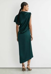 Pikk, keha järgnev tume roheline kleit lühikeste, drapeeritud varrukatega ja asümmeetrilise äärega. Valmistatud siledast kangast, kantud koos rihmikestega kõrgete kontsadega.