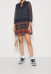 Marinblå klänning med genomskinliga långa ärmar, blommönster, volangkrage och mönstrad nederkant; i kombination med vita strumpor och marinblå sneakers.