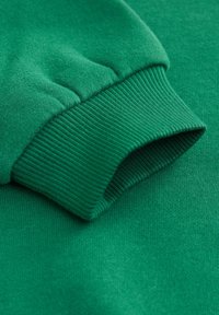 Zöld pulóver anyag lágy tapintással. Bordázott mandzsettával és sima anyaggal, közeli kép a varrás részleteiről.