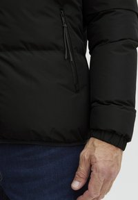 Črna puffer jakna z gladkim, mat zaključkom, ki vključuje žepek z zadrgo na strani in rebrasto obrobo na rokavih. Oblečena z modrimi koli.