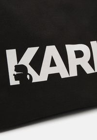 Czarna torba na ramię z płótna z odważnym, białym napisem "KARL" i sylwetką, wskazującym na element projektu. Gładka tekstura.
