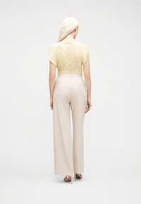 Γυναίκα με μακριά ξανθά μαλλιά, που φοράει κίτρινη μπλούζα με ανοιχτά σχέδια, μπεζ παντελόνια φαρδιάς γραμμής και μπεζ ψηλοτάκουνα σανδάλια, να στέκεται με την πλάτη προς τα εμπρός.