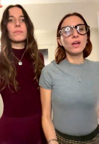 Two young women standing indoors; one with long brown hair in a maroon top, the other with red hair, glasses, and a gray t-shirt.