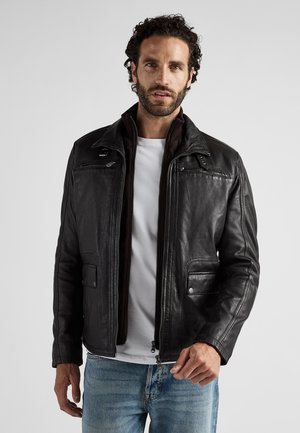 Mand iført en sort læderjakke over en mørk lynlås-trøje og en hvid t-shirt, parret med lyseblå jeans, stående mod en hvid baggrund.