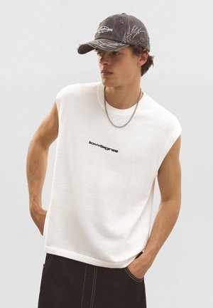 Mladý muž nosiaci biely pletený top bez rukávov s čiernym logom, čiernu opotrebovanú čiapku, strieborný retiazkový náhrdelník a čierne nohavice s bielym šitím.