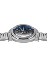 Silverfärgad rostfri klocka med en blå solstråledial, skelettdesign och silverfärgade timmarkörer. Har ett länkarmband och en oktagonal klockkropp.