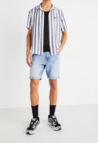 Hombre con camisa de manga corta a rayas verticales en azul y blanco, camiseta negra, pantalones cortos de mezclilla azul claros, calcetines negros y zapatillas negras y blancas.
