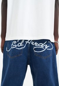 Džínové kalhoty s vyšívaným nápisem "Ed Hardy" v světlemodré barvě na zadních kapsách. Bílé bavlněné tričko s mírně zakřiveným lemem nad džínami.