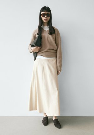 Donna che indossa un maglione marrone oversize, gonna lunga beige, scarpe slip-on scure, grandi occhiali da sole, tiene una pochette nera con trama contro una parete semplice.