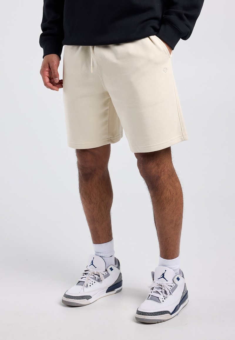 Personne portant un short beige jusqu'aux genoux, des baskets blanches et grises avec des logos bleus, et un haut à manches longues noir, debout sur un fond blanc.