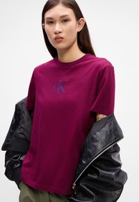 Μαρόνος βαμβακερός μπλουζάκι με στρογγυλή λαιμόκοψη, κοντά μανίκια και ένα μικρό, μπλε λογότυπο Calvin Klein στο στήθος. Φορεμένο κάτω από μια μαύρη δερμάτινη ζακέτα.