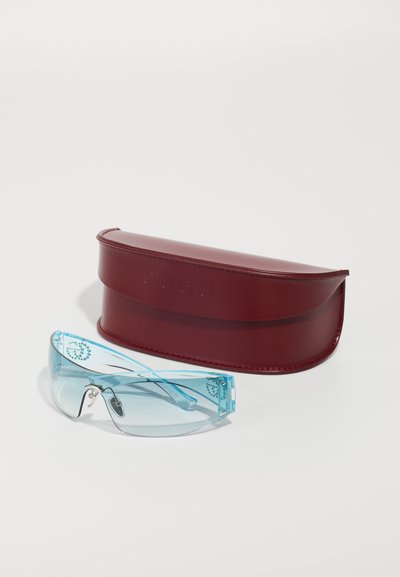Mėlyna atspalvio apvalūs saulės akiniai su akmenukų detalėmis šalia raudonos odinės Gucci akinių dėklo ant balto fono.