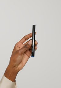 Stylo de maquillage noir à double extrémité avec une finition mate ; une extrémité est bleue et l'autre est destinée à l'application. Tenue entre les doigts contre un fond clair.